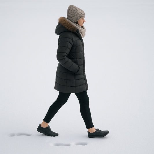 En kvinna i vinterkläder som går på snö med minimalistiska barfotaskor, tydligt synliga tåspår i snön. Alt: barfotaskor dam vinterfotografering